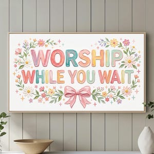May include: A framed art print with the words "WORSHIP WHILE YOU WAIT" in colorful, bubble letters. The text is surrounded by a floral wreath and a pink bow. The artwork is displayed above a white cabinet with decorative vases and a bowl.