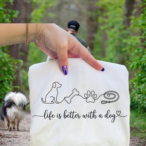 May include: A white tote bag featuring a black line drawing of a dog, bone, paw print, and leash. The text "life is better with a dog" is written below the drawing. The bag is held by a person in a park, with a dog on a lead.