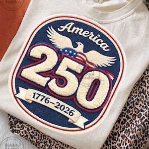 Puede incluir: Camiseta de manga larga color crema con un parche grande con la palabra "America", el número "250" y los años "1776-2026". El parche incluye un águila, la bandera estadounidense y un borde azul y rojo. La camiseta se combina con pantalones con estampado de leopardo.