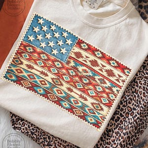 May include: Cream-colored t-shirt with a large, embroidered American flag design. The flag features a blue field with white stars and a patterned design in red, brown, and turquoise. Leopard print pants are visible below the shirt. The shirt has the text "COMFORT COLORS" on the tag.