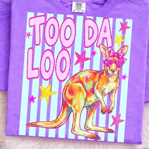 Op de afbeelding: Paarse t-shirt met een kleurrijke kangoeroe-afbeelding. De kangoeroe draagt een hartvormige zonnebril en een strik. De tekst "TOO DA LOO" is roze, met roze en gele sterren. De kangoeroe is in tinten van oranje, geel en roze.