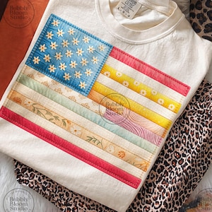 Könnte beinhalten: Cremefarbenes T-Shirt mit einem Patchwork-Design der amerikanischen Flagge. Die Flagge hat ein blaues Quadrat mit gestickten weißen Blumen und bunten Streifen. Das Shirt wird mit einer Hose mit Leopardenmuster kombiniert. Der Text "COMFORT COLORS" ist sichtbar.