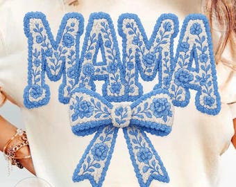 Puede incluir: Camiseta color crema con la palabra "MAMA" bordada en azul floral y un diseño de lazo a juego. El diseño tiene una apariencia texturizada y tridimensional. Esta camiseta informal es una prenda elegante.