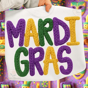 May include: White sweatshirt with the words "MARDI GRAS" in large, textured, colorful letters. The letters are purple, yellow, and green. The sweatshirt is held up by a person's hand. The background has crochet decorations.