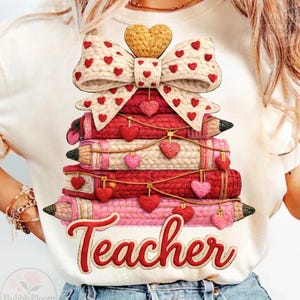 May include: Cream-colored t-shirt featuring a stack of books designed as a Christmas tree, adorned with a bow and hearts. The word "Teacher" is written in red script. The design includes pencils and decorative elements.