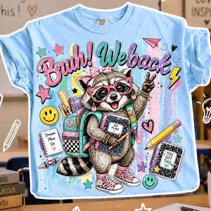Puede incluir: Camiseta azul claro con un gr&aacute;fico de mapache con gafas y una mochila, sosteniendo un cuaderno. La camiseta tiene el texto "Bruh! Weback" y gr&aacute;ficos escolares. La camiseta est&aacute; sobre una mesa de madera.