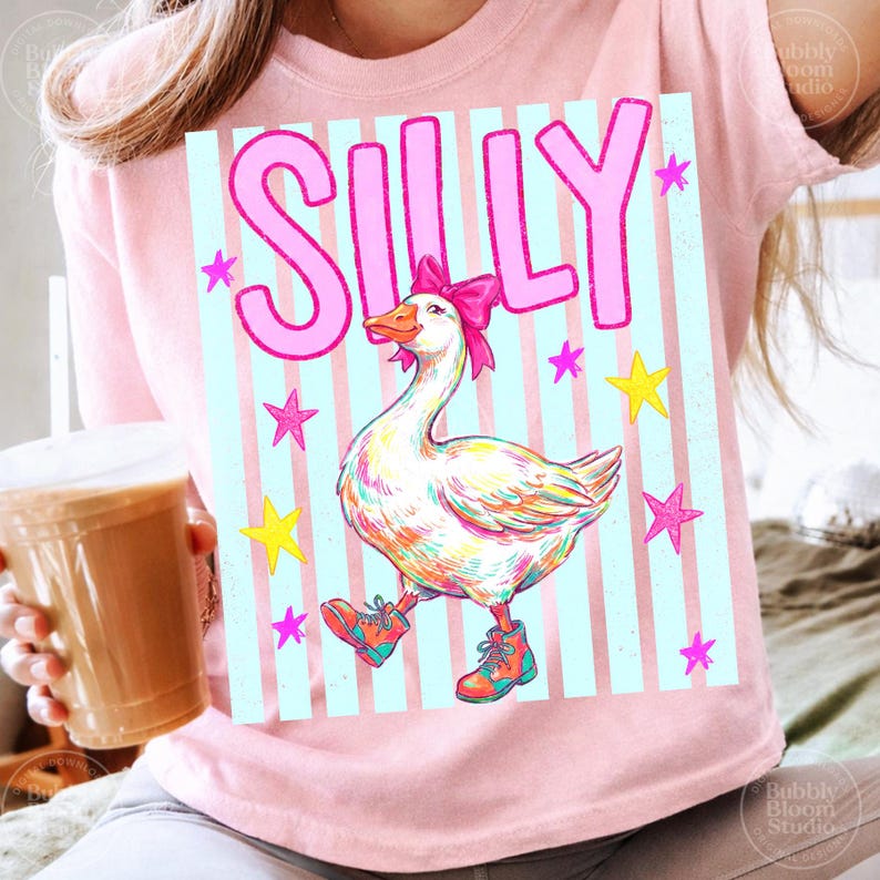 Puede incluir: Camiseta rosa claro con un dibujo de un ganso con botas y un lazo rosa. La palabra "SILLY" est&aacute; en rosa encima del ganso, con estrellas rosas y amarillas. El fondo tiene rayas verticales azul claro.