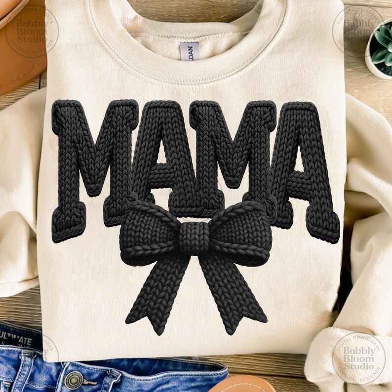 May include: Cream-colored sweatshirt with the word "MAMA" in large, black, knitted letters. A black knitted bow is centered below the word. The sweatshirt is a casual, comfortable style.