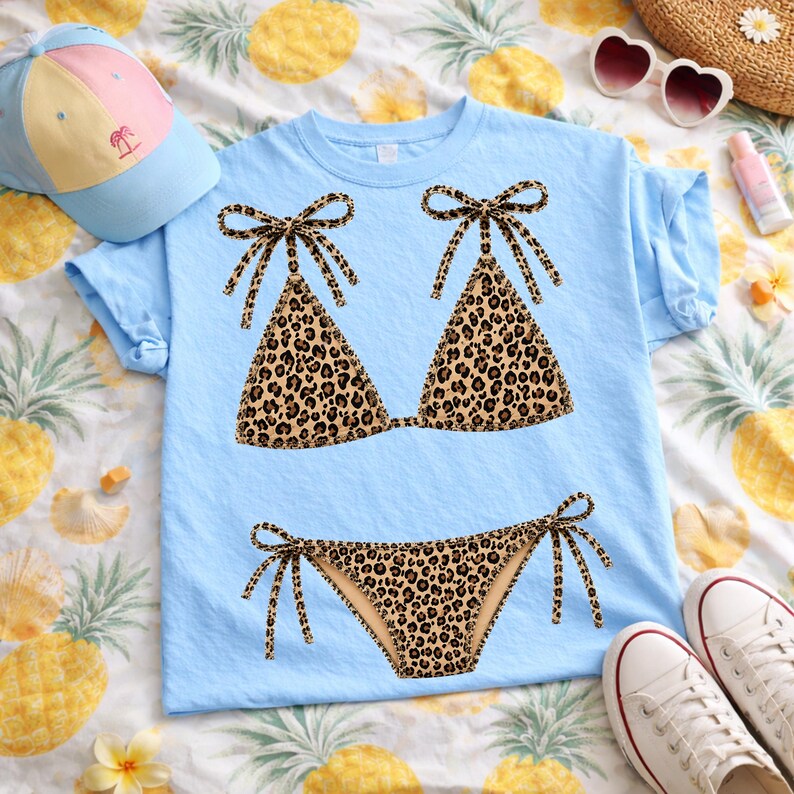 May include: Light blue t-shirt featuring a leopard print bikini design. The bikini top and bottom are brown with black spots and tied with brown bows. A light blue cap, heart-shaped sunglasses, and white sneakers are also visible.