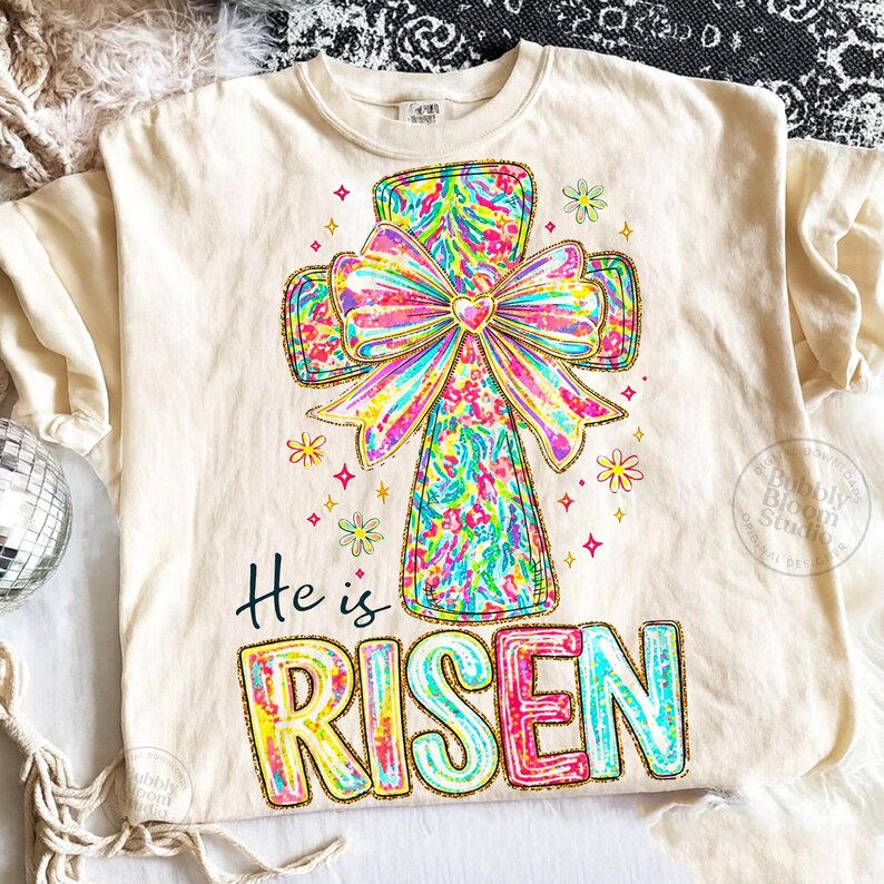 May include: Cream-colored t-shirt featuring a colorful cross design with a large bow and the words "He is Risen." The cross and text have a glittery outline. The design includes floral and star accents, creating a festive and celebratory look.