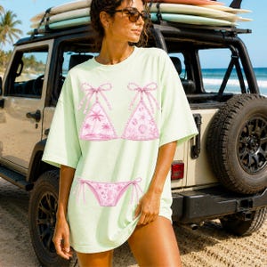 May include: A light green t-shirt featuring a pink bikini design with palm trees, seashells, and bows. The t-shirt is worn by a person standing near a beige Jeep with surfboards on top. The background includes a beach and ocean.