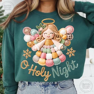 May include: A teal green sweatshirt featuring a crocheted angel design with a halo and colorful wings. The angel is surrounded by snowflakes and the words "O Holy Night" in a script font. The design is a digital download.