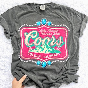 May include: Dark gray t-shirt featuring a distressed pink and teal graphic with the text "Coors" and "Rocky Mountain Mountain Water." The design includes a mountain scene and is framed with faux jewels and pearls. The text "Golden, Colorado" is also included.