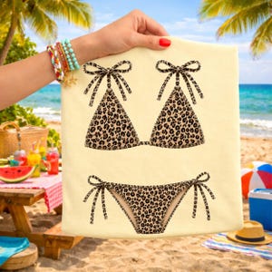 May include: A beige beach towel featuring a leopard print bikini top and bottom. The bikini top has bow ties at the top, and the bikini bottom has bow ties on the sides. The towel is held up on a beach with palm trees and the ocean in the background.