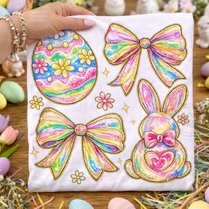 May include: White fabric featuring colorful Easter-themed designs. The design includes a decorated egg, two bows, a bunny with a heart, and flowers. The illustrations have rainbow colors with gold glitter accents, perfect for Easter.
