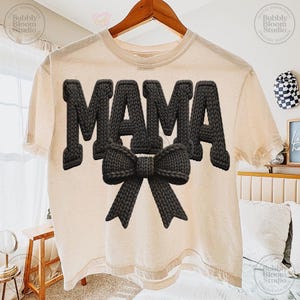May include: A cream-colored t-shirt with the word "MAMA" in large, textured, black letters and a black bow. The shirt is hanging on a wooden hanger.