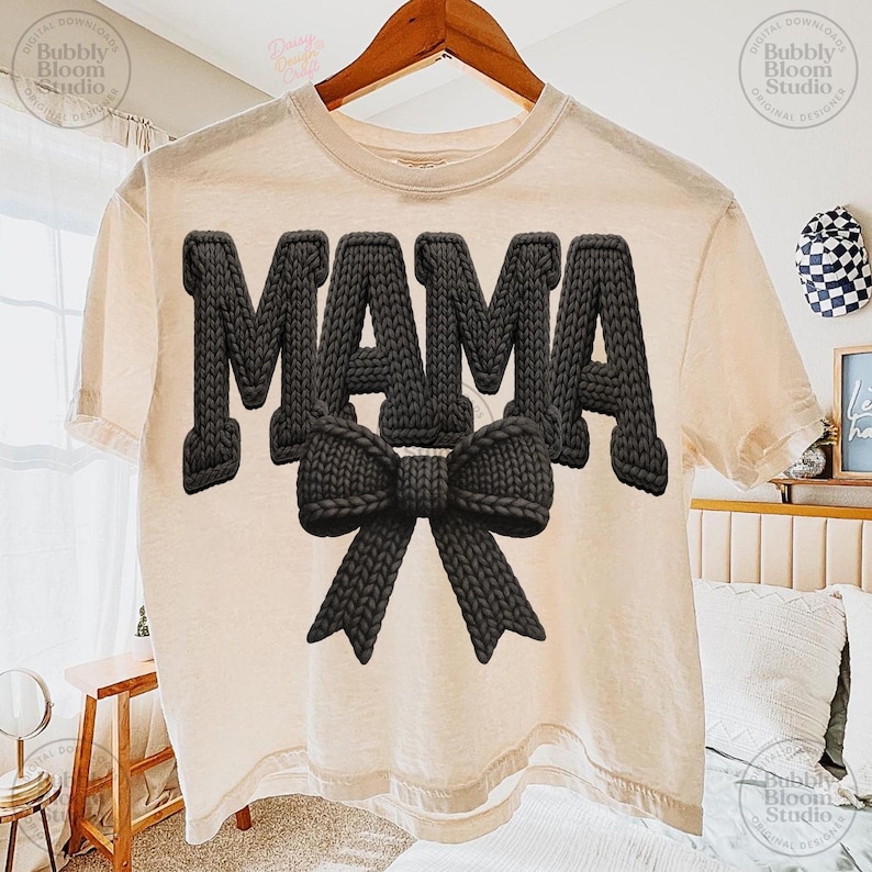 May include: A cream-colored t-shirt with the word "MAMA" in large, textured, black letters and a black bow. The shirt is hanging on a wooden hanger.