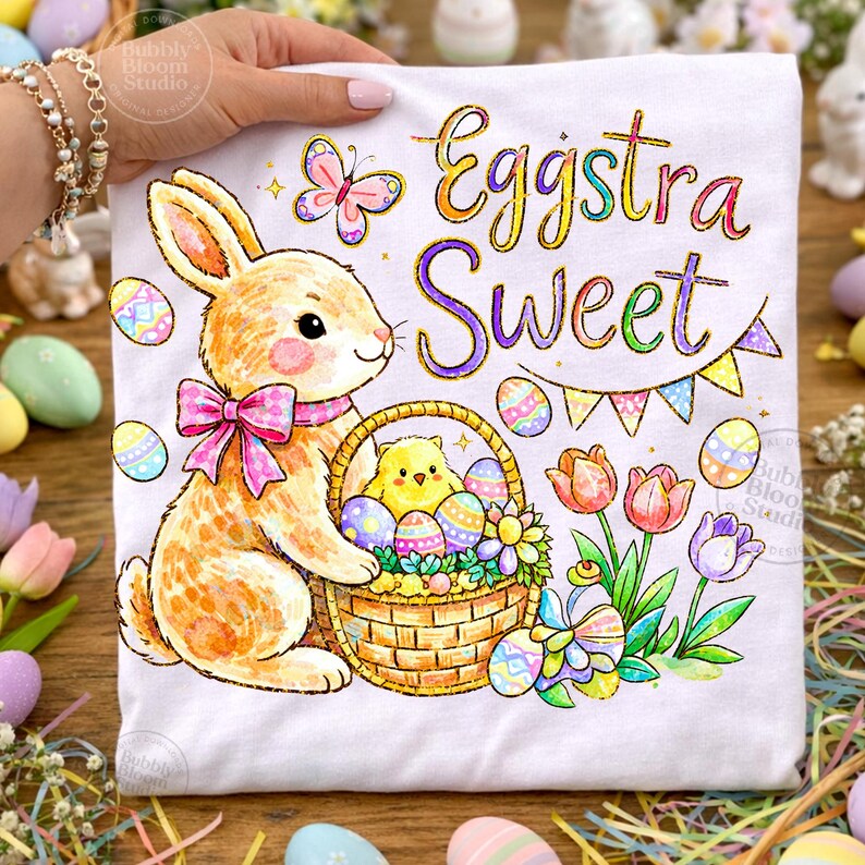 May include: White t-shirt with an Easter-themed design. It features a cartoon bunny with a pink bow, a basket of eggs, and a chick. The words "Eggstra Sweet" are written in colorful script, with a butterfly and flowers.