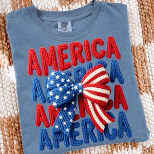 May include: A blue t-shirt with the word "AMERICA" in red, white, and blue knitted letters. A bow with a star-spangled blue section and red and white stripes is centered on the shirt. The shirt is a Comfort Colors brand.