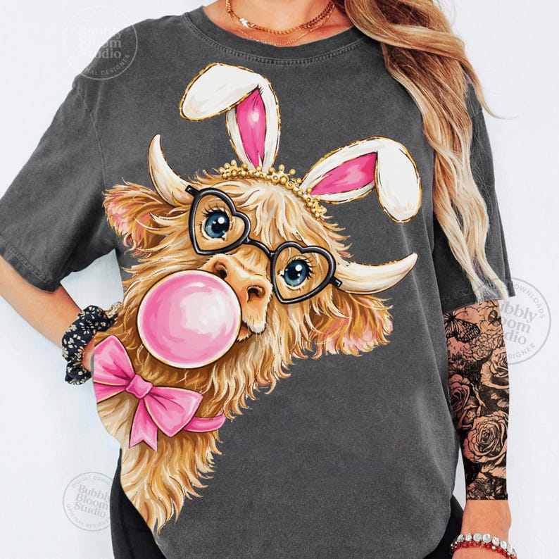 May include: Dark gray t-shirt featuring a Highland cow with bunny ears, heart-shaped glasses, and a pink bubblegum bubble. The cow has a pink bow and a gold crown. The design is whimsical and fun.