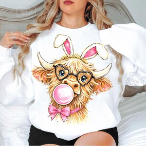 May include: White sweatshirt featuring a cartoon Highland cow wearing heart-shaped glasses, bunny ears, and a pink bow. The cow is blowing a pink bubblegum bubble. The design has a gold crown and is perfect for Easter.