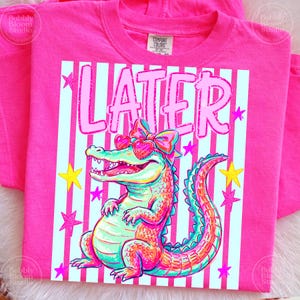 Puede incluir: Una camiseta rosa brillante con un caim&aacute;n colorido con gafas de sol en forma de coraz&oacute;n y un lazo. La palabra "LATER" est&aacute; impresa encima del caim&aacute;n sobre un fondo a rayas con estrellas. La camiseta es de material suave.
