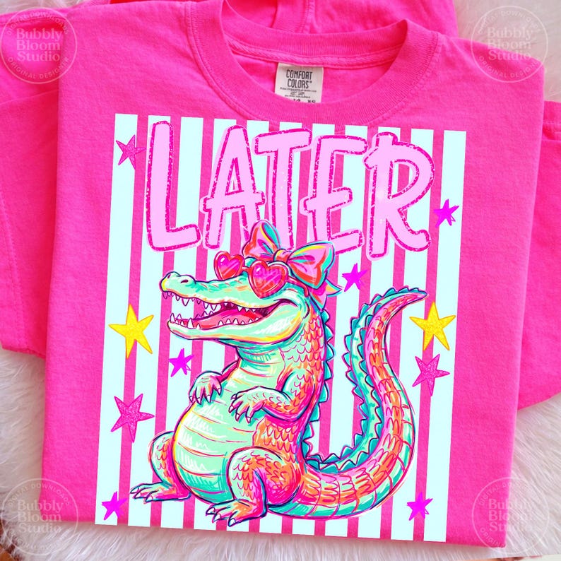 Puede incluir: Una camiseta rosa brillante con un caim&aacute;n colorido con gafas de sol en forma de coraz&oacute;n y un lazo. La palabra "LATER" est&aacute; impresa encima del caim&aacute;n sobre un fondo a rayas con estrellas. La camiseta es de material suave.