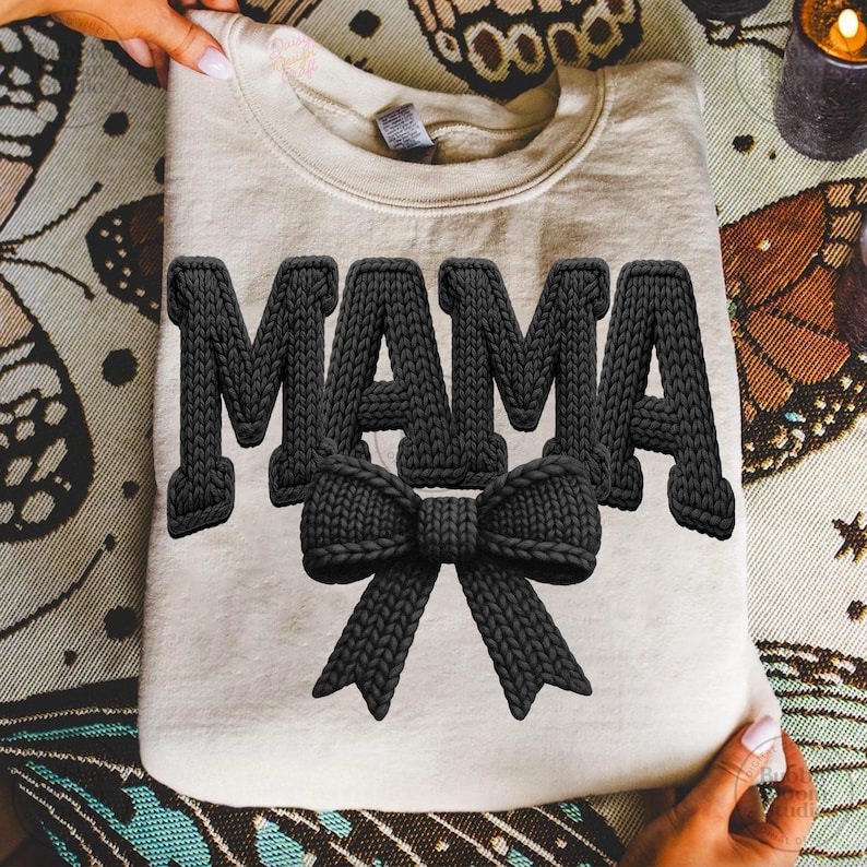 May include: A beige sweatshirt with the word "MAMA" in large, black, textured letters. A black bow is centered below the word. The sweatshirt is held up by hands, and the background is a patterned fabric.