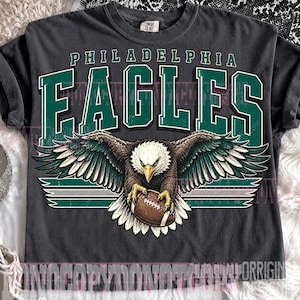 May include: A charcoal gray short-sleeve t-shirt featuring a graphic print of a bald eagle with wings spread, clutching a brown football. The text PHILADELPHIA EAGLES is printed in large green and white block letters above the eagle illustration.