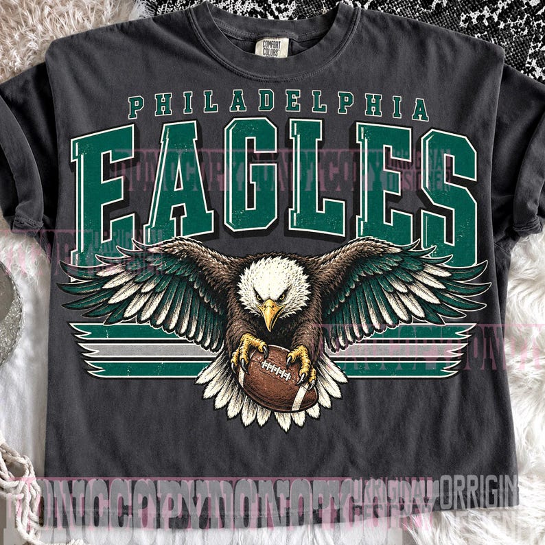 May include: A charcoal gray short-sleeve t-shirt featuring a graphic print of a bald eagle with wings spread, clutching a brown football. The text PHILADELPHIA EAGLES is printed in large green and white block letters above the eagle illustration.