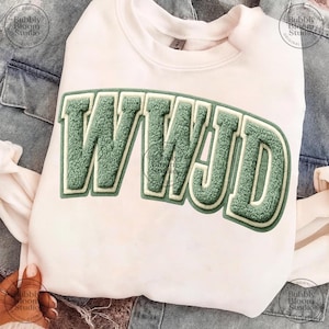 May include: A white sweatshirt with the letters "WWJD" in a green, textured, collegiate-style font. The sweatshirt is displayed on a denim background.