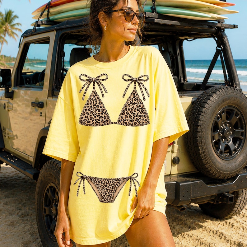 May include: Yellow oversized t-shirt featuring a leopard print bikini design. The bikini top and bottom are printed on the front of the shirt. The shirt is worn by a person standing near a beach and a vehicle with surfboards on top.