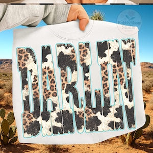 May include: White sweatshirt with the word "DARLIN" in large, distressed lettering. The letters feature a cow print and leopard print design with a turquoise outline. The background shows a desert landscape with cacti and mountains.