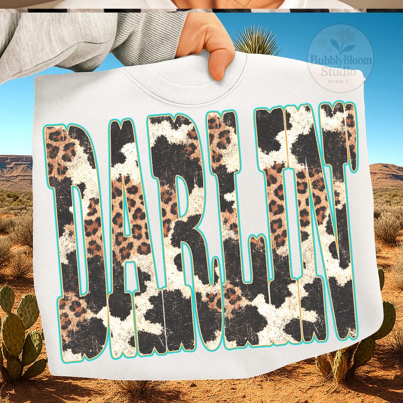 May include: White sweatshirt with the word "DARLIN" in large, distressed lettering. The letters feature a cow print and leopard print design with a turquoise outline. The background shows a desert landscape with cacti and mountains.