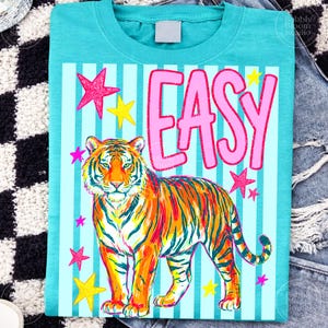 Puede incluir: Camiseta turquesa con una colorida ilustraci&oacute;n de tigre y la palabra "EASY" en rosa. El dise&ntilde;o incluye estrellas rosas y amarillas sobre un fondo a rayas azul claro.