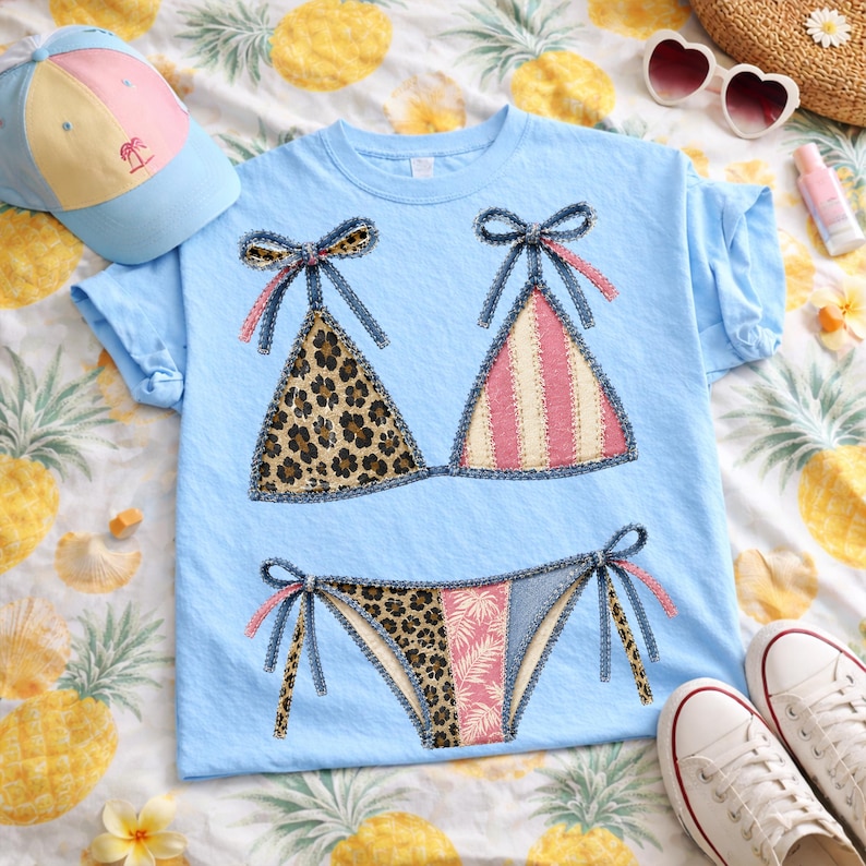 May include: Light blue t-shirt featuring a bikini design with leopard print and pink striped patterns. The bikini top and bottom are adorned with denim-like bows. A light blue cap, heart-shaped sunglasses, and white sneakers are also visible.