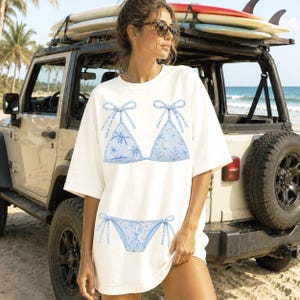 May include: White t-shirt featuring a light blue bikini design. The bikini top and bottom are adorned with a tropical pattern and bow details. The image is taken outdoors with a Jeep and surfboards in the background.