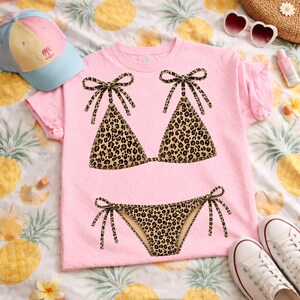 May include: Pink t-shirt featuring a leopard print bikini design. The bikini top and bottom are detailed with brown and black spots and tied with bows. A light blue and pink baseball cap and white sneakers are also in the image.