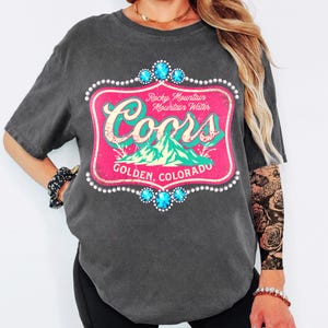 Peut inclure: T-shirt gris fonc&eacute; avec un motif rose et turquoise. Le motif indique "Coors Rocky Mountain Mountain Water Golden, Colorado" et est encadr&eacute; de perles blanches et de gemmes bleues.