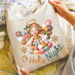 May include: Cream-colored sweatshirt featuring an embroidered angel design with a halo, wings, and a dress made of colorful yarn. The text "O Holy Night" is embroidered below the angel, along with gold snowflakes.