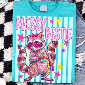 Puede incluir: Camiseta turquesa con un dise&ntilde;o de mapache colorido con gafas de sol en forma de coraz&oacute;n y una bebida. El texto "MESSY BESTIE" es rosa, con estrellas rosas y amarillas. Fondo a rayas.
