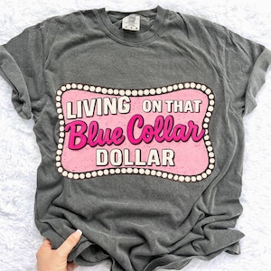 May include: Dark gray t-shirt with a pink and white graphic that reads "LIVING ON THAT Blue Collar DOLLAR." The graphic is framed with a white dotted border. The shirt has short sleeves and a crew neck.