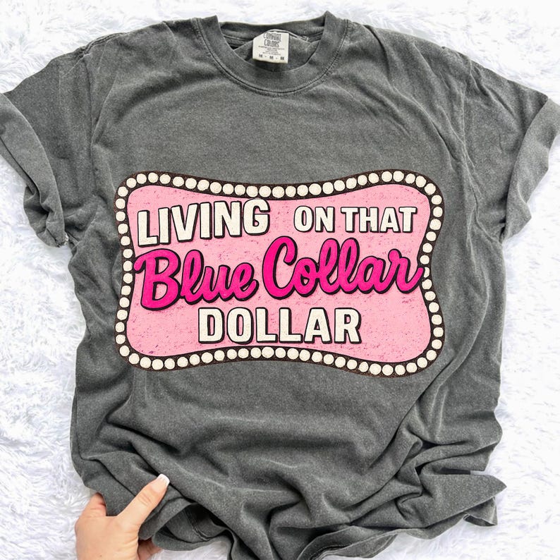 May include: Dark gray t-shirt with a pink and white graphic that reads "LIVING ON THAT Blue Collar DOLLAR." The graphic is framed with a white dotted border. The shirt has short sleeves and a crew neck.