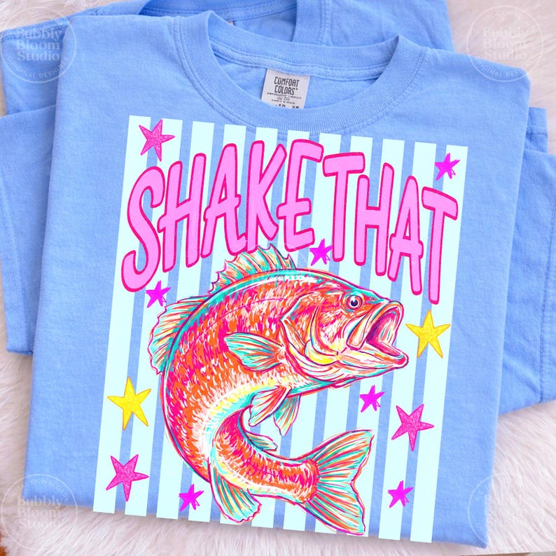Puede incluir: Camiseta azul claro con la frase "SHAKE THAT". El dise&ntilde;o presenta un pez colorido y estrellas. La camiseta est&aacute; doblada.