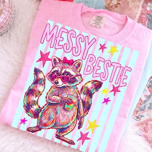 Puede incluir: Camiseta rosa con un gr&aacute;fico de un mapache con gafas de sol en forma de coraz&oacute;n y un lazo, sosteniendo una bebida. El texto "MESSY BESTIE" est&aacute; encima del mapache, con estrellas. La camiseta tiene una etiqueta Comfort Colors.