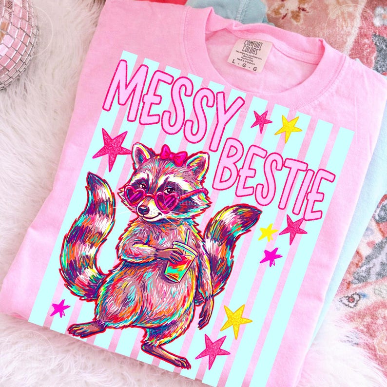 Puede incluir: Camiseta rosa con un gr&aacute;fico de un mapache con gafas de sol en forma de coraz&oacute;n y un lazo, sosteniendo una bebida. El texto "MESSY BESTIE" est&aacute; encima del mapache, con estrellas. La camiseta tiene una etiqueta Comfort Colors.