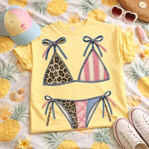 May include: Yellow t-shirt with a bikini design. The bikini top features leopard print and pink stripes, with denim-like bows. The matching bikini bottom has leopard print and a pink floral pattern. A light blue and pink cap and white sneakers are also visible.