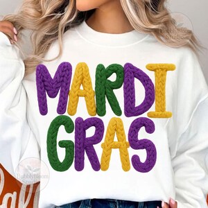 May include: White sweatshirt with the words "MARDI GRAS" in large, textured letters. The letters are in purple, yellow, and green, with a knitted appearance. The design is centered on the front of the shirt.
