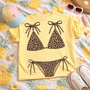 May include: Yellow t-shirt featuring a leopard print bikini design. The bikini top and bottom are detailed with black bows. A light blue and pink cap, white sneakers, and sunglasses are also visible, suggesting a summer theme.