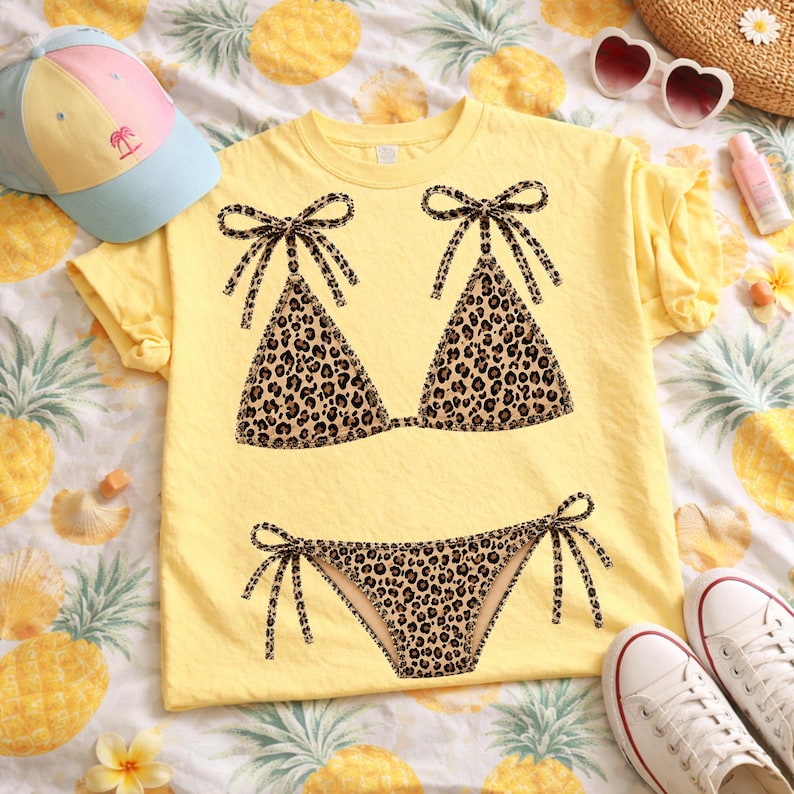 May include: Yellow t-shirt featuring a leopard print bikini design. The bikini top and bottom are detailed with black bows. A light blue and pink cap, white sneakers, and sunglasses are also visible, suggesting a summer theme.